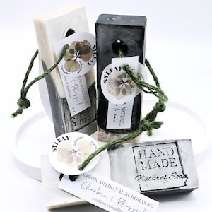 Puede incluir: Barras de jabón natural artesanal en varias formas y colores, algunas con etiquetas y cuerda verde. Una barra es negra, otra blanca con una franja oscura y una tercera gris con las palabras "HAND MADE Natural Soap". Las barras de jabón están etiquetadas con las palabras "SAVON ARTISANAL SURGRAS 8% Charbon & Rhassoul".