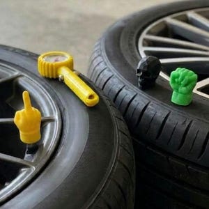 May include: A yellow tire valve cap shaped like a hand with a middle finger extended, a yellow tire pressure gauge, a black skull-shaped valve cap, and a green fist-shaped valve cap, all positioned on two black rubber tires with metal rims.