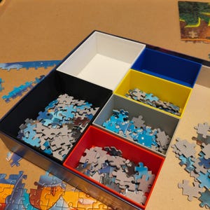 Könnte beinhalten: Ein mehrfarbiger Puzzlesortierer mit sechs Fächern in verschiedenen Farben: Schwarz, Weiß, Blau, Gelb, Grau und Rot. Die Fächer sind mit Puzzleteilen in Blau-, Grau- und Weißtönen gefüllt. Der Sortierer steht auf einer Holzoberfläche.