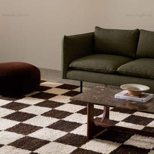 Puede incluir: Escena de salón con una alfombra a cuadros marrón y crema. Se ve un sofá verde oscuro y una mesa de centro de mármol marrón con un libro y un cuenco. Un puf marrón está a la izquierda.