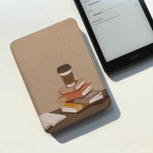May include: A tan e-reader case featuring a graphic illustration of a stack of books with a coffee cup on top. The cup has a heart design. An open book lies at the base of the stack. A black Kindle e-reader with a settings menu is visible nearby.