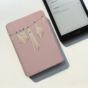 以下が含まれることがあります： くすんだピンク色の電子書籍リーダーケース。濃い赤色の物干しロープに緑色の洗濯バサミで吊るされた、5体のオフホワイトの幽霊のグラフィックイラストが描かれています。幽霊はそれぞれ大きさが異なり、シンプルな黒い目が付いています。