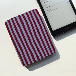 Puede incluir: Un lector de libros electrónicos Kindle y una funda. La funda tiene un patrón de rayas verticales alternas en burdeos y azul claro. El Kindle negro muestra la configuración en su pantalla.