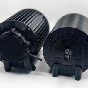 May include: Two black cylindrical objects with detailed designs. One has ribbed sides, the other a gear-like pattern. Both have small legs for support. The objects appear to be 3D printed. Measurements are in cm.