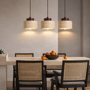 May include: Three cylindrical pendant lights with textured beige stone shades and dark brown metal hardware hang above a dining table. The table features a bowl of oranges and chairs with black frames and woven cane backrests.