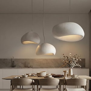 Peut inclure: Trois lampes suspendues blanches modernes, de forme organique et lisse, sont suspendues au-dessus d'une table à manger. Les lampes émettent une lueur chaleureuse de l'intérieur. Les lampes sont suspendues au plafond par de fins câbles.
