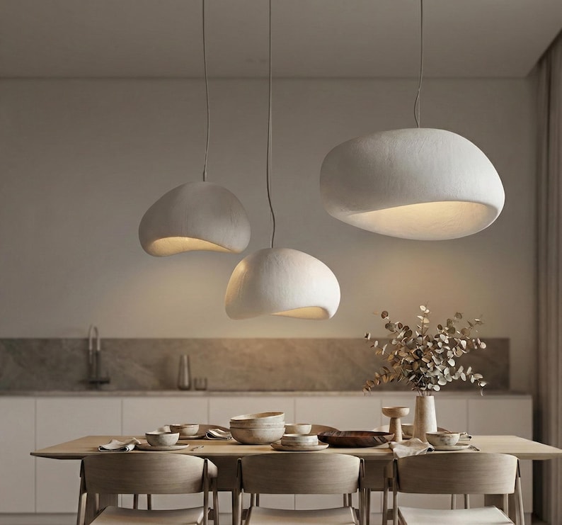 May include: Three modern, white pendant lights with a smooth, organic shape hang above a dining table. The lights have a warm, inviting glow from the interior. The lights are suspended from the ceiling by thin cords.