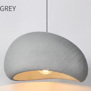 May include: A grey, textured pendant light with a curved, organic shape. The light fixture has a warm, golden interior and hangs from a single cord. The text "B - GREY" is visible in the upper left corner.