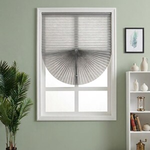 May include: A grey pleated window shade with a semi-circular fan-like bottom section, installed within a white window frame. The shade features horizontal lines and a central pull cord, partially covering the glass panes of the window.