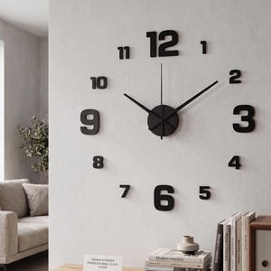 Peut inclure: Une horloge murale sans cadre composée de grands chiffres noirs en gras, allant de un à douze, disposés en cercle. L'horloge présente un moyeu central circulaire noir avec deux fines aiguilles noires indiquant l'heure sur le mur.