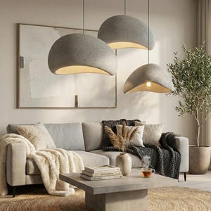 May include: Three gray, dome-shaped pendant lights hang above a living room. The lights have a textured surface and a warm, inviting glow from the exposed bulbs. The room features a light-colored sofa, a coffee table, and a decorative olive tree.