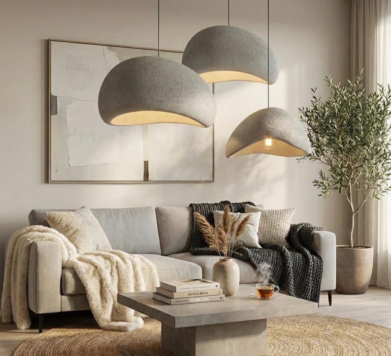 May include: Three gray, dome-shaped pendant lights hang above a living room. The lights have a textured surface and a warm, inviting glow from the exposed bulbs. The room features a light-colored sofa, a coffee table, and a decorative olive tree.