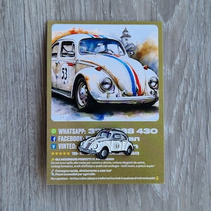 Può includere: Una cartolina con un dipinto ad acquerello di un'auto d'epoca bianca con strisce da corsa e il numero 53. La cartolina presenta anche una piccola spilla a forma di auto con lo stesso design.