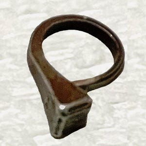 May include: A weathered, bronze-coloured ring with a distinctive shape. The ring features a rounded top and a rectangular base, with visible texture and a slightly aged appearance. The design suggests a vintage or antique style.