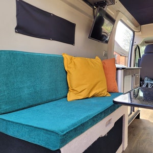 May include: A teal-colored bench seat with two pillows, one yellow and one orange, in a camper van. The bench seat is in front of a black table with a black and white speckled surface.