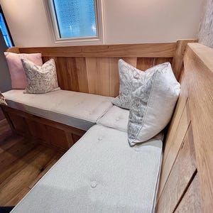 A kitchen dining banquette seating area with a set of made to measure foam bench cushions. The storage seating area is made up with light brown wood panelling and it has three piped and buttoned bench seating cushions on top made with an oatmeal, cream coloured fabric. The hardwearing twill fabric bench cushion cover has a stain resistant treatment making this box edged bench cushion perfect for use around kids, children and pets. Hardwearing and easy clean seat cushions for benches and seats.