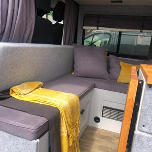 The interior of a renovated camper van with dark grey firm foam bench cushions inside. The converted campervan has comfortable thick upholstery foam cushions held in place with velcro strips on the underside of the cushions. The motor home bed cushions pull out to form a sleeping area with memory foam for comfort. The stain resistant bench cushion covers for the camper van conversion are made to measure for a plywood camper van build. Cut outs to fit around the van wall shaping for a perfect fit