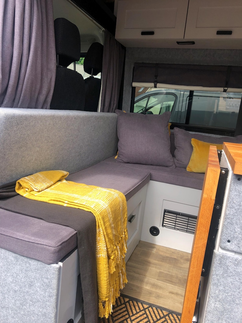 The interior of a renovated camper van with dark grey firm foam bench cushions inside. The converted campervan has comfortable thick upholstery foam cushions held in place with velcro strips on the underside of the cushions. The motor home bed cushions pull out to form a sleeping area with memory foam for comfort. The stain resistant bench cushion covers for the camper van conversion are made to measure for a plywood camper van build. Cut outs to fit around the van wall shaping for a perfect fit