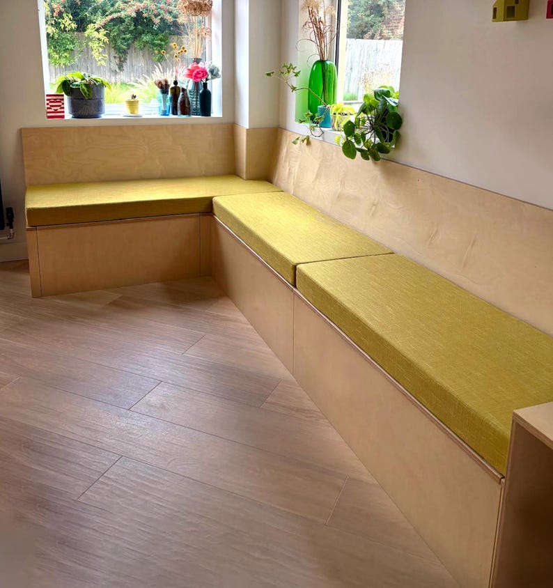 A bright kitchen banquette seating area build in wooden storage cupboard lift up lid seat. Wooden flooring kitchen. Yellow indoor bench cushions made to fit the plywood seating bench. Thick firm foam supportive home office seating cushions made to measure. Clean, unpiped bench cushions wipe clean, machine washable bench cushion covers, pet friendly. Bespoke high quality long lasting bench cushion kept in place with anti-slip cloth non permanent fixing. Welt free box edge seat bad & back cushion