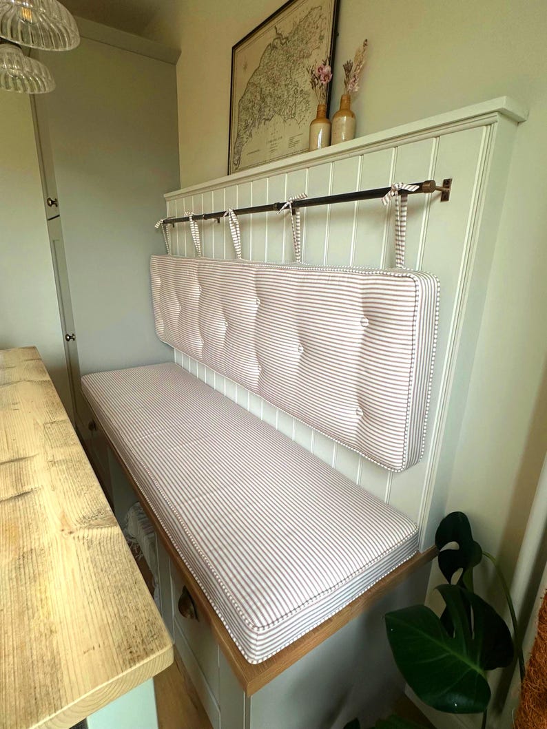 A light sage green painted dining banquette booth seating area in Farrow and Ball, Little Greene paint. The storage bench seat has a custom made indoor bench cushion with firm foam and dacron wadding inside. The bench cushion cover is hand made in the UK with a 100% cotton french country chic ticking fabric from Ian Mankin in a light beige colour ticking stripe. The back cushion is hung from a metal bar mounted to the wall with fabric ties and is button tufted for extra texture. Stain resistant