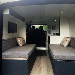 Inside a refurbished camper van - wood panelled flooring and walls in light cream paint. A pull out wooden bed base with bench seat cushions that form a  mattress when put together. Firm fire safe upholstery foam inside for support with machine washable dark grey woven linen look fabric covers. Durable and long lasting seat cushions with back bolster cushions in the same grey cushion fabric. Stowaway beds in a camper van conversion with made to measure cushions high quality and long lasting.