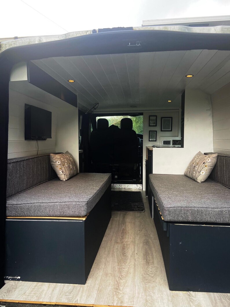 Inside a refurbished camper van - wood panelled flooring and walls in light cream paint. A pull out wooden bed base with bench seat cushions that form a  mattress when put together. Firm fire safe upholstery foam inside for support with machine washable dark grey woven linen look fabric covers. Durable and long lasting seat cushions with back bolster cushions in the same grey cushion fabric. Stowaway beds in a camper van conversion with made to measure cushions high quality and long lasting.