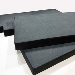 May include: Three rectangular, dark gray cushions with black borders are stacked at different angles. The cushions have a textured fabric appearance and are placed against a white background. The cushions appear to be for seating.