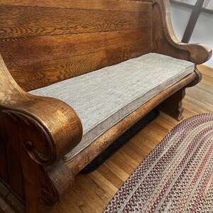 A thick, heavy looking oak wood bench with sturdy arms. A thin bench cushion on top of the church pew in a light brown herringbone patterned upholstery fabric with firm foam inside. Made to measure indoor bench cushion in patterned fabrics with stain resistant treatment. Pet friendly bench cushion, wipe clean window seat cushion with piped edges. Zippered bench cushion cover for dining banquette. Hanging back cushions. Overlocked finished seams, Ercol sofa cushions shaped edges, curved corners