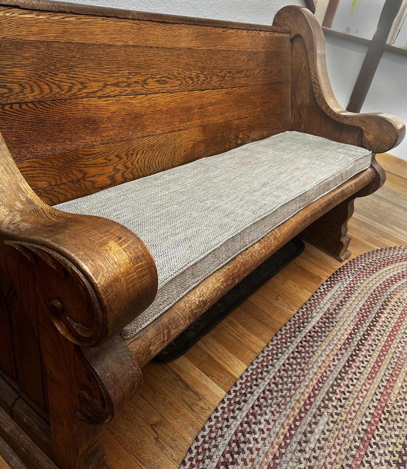 A thick, heavy looking oak wood bench with sturdy arms. A thin bench cushion on top of the church pew in a light brown herringbone patterned upholstery fabric with firm foam inside. Made to measure indoor bench cushion in patterned fabrics with stain resistant treatment. Pet friendly bench cushion, wipe clean window seat cushion with piped edges. Zippered bench cushion cover for dining banquette. Hanging back cushions. Overlocked finished seams, Ercol sofa cushions shaped edges, curved corners