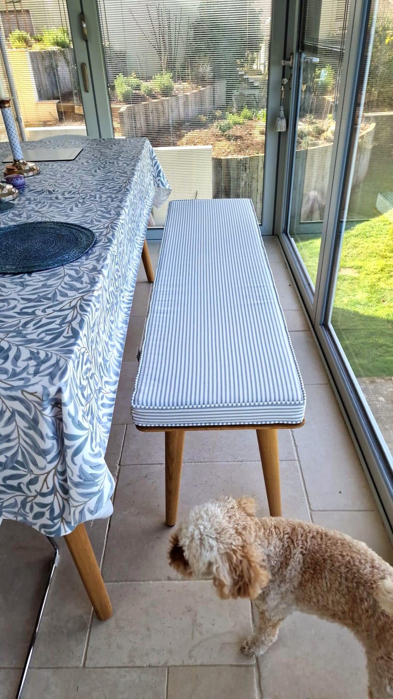 A bright, sunny conservatory dining room with a wooden table and bench seat. The table has a william morris white and blue design table cloth. The wooden seating bench has a bespoke made bench cushion in a white and blue cotton ticking fabric. The french style chic ticking fabric has blue and white stripes running down the length of the cushion. The foam bench cushion has sturdy velcro straps to keep the cushion in place when sat on and the cushion cover has a zip at one side for removal.