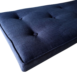 May include: A rectangular, navy blue cushion with button tufting. The cushion has a tailored edge and is made of a textured fabric. The design is simple and modern, suitable for a bench or seating area.