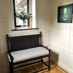 A classic wood panelled room corner painted in an off white Farrow and Ball colour. There is a dark wooden church pew bench that has a well made french ticking fabric bench cushion with cut outs for the bench arms. The cushion is a cotton fabric in a black and white ticking and has piped edges and a zip at the back of the cover. The custom shaped pew cushion has thin firm foam insert and piping at the edges. The stain resistant, striped fabric is a 100% cotton upholstery fabric made in the UK