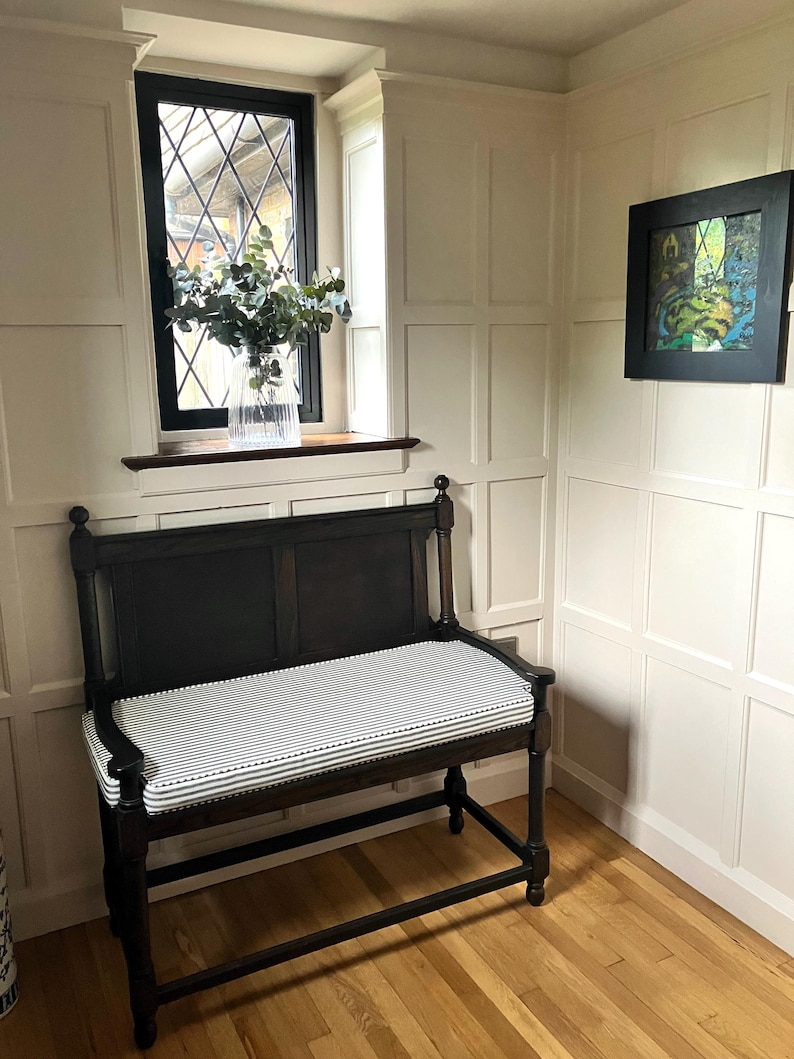 A classic wood panelled room corner painted in an off white Farrow and Ball colour. There is a dark wooden church pew bench that has a well made french ticking fabric bench cushion with cut outs for the bench arms. The cushion is a cotton fabric in a black and white ticking and has piped edges and a zip at the back of the cover. The custom shaped pew cushion has thin firm foam insert and piping at the edges. The stain resistant, striped fabric is a 100% cotton upholstery fabric made in the UK