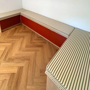 A U shaped storage cupboard seating area for a dining room. Stain resistant custom made bench cushions in an olive green and oatmeal coloured candy stripe pattern. Cotton natural fabric bench cushion covers made in the UK. Ian Mankin ticking stripe fabric with piping at the edges of the box edge cushions in the same green and white stripe pattern. Firm foam inside with velcro strips underneath the cushions. Pull out cupboard storage unit for dining room seating. Bespoke bench cushions with zips
