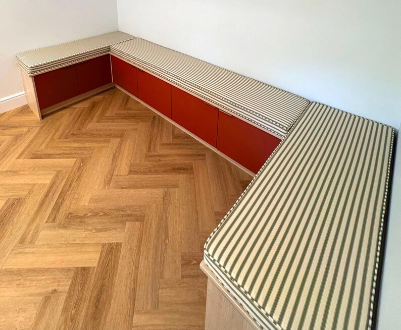 A U shaped storage cupboard seating area for a dining room. Stain resistant custom made bench cushions in an olive green and oatmeal coloured candy stripe pattern. Cotton natural fabric bench cushion covers made in the UK. Ian Mankin ticking stripe fabric with piping at the edges of the box edge cushions in the same green and white stripe pattern. Firm foam inside with velcro strips underneath the cushions. Pull out cupboard storage unit for dining room seating. Bespoke bench cushions with zips
