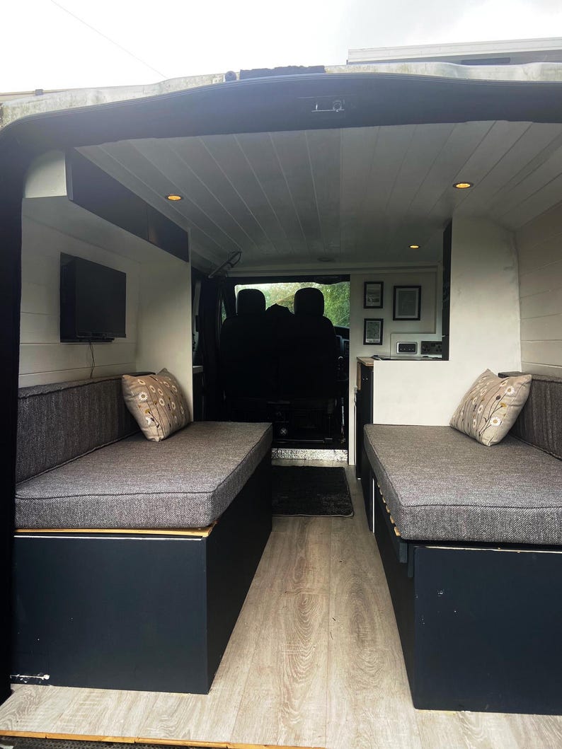 Inside a refurbished camper van - wood panelled flooring and walls in light cream paint. A pull out wooden bed base with bench seat cushions that form a  mattress when put together. Firm fire safe upholstery foam inside for support with machine washable dark grey woven linen look fabric covers. Durable and long lasting seat cushions with back bolster cushions in the same grey cushion fabric. Stowaway beds in a camper van conversion with made to measure cushions high quality and long lasting.