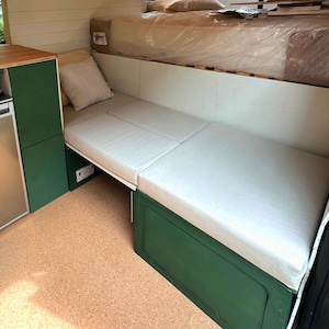 May include: Interior view of a camper van with a built-in bed and seating area. The seating area has a light gray cushion and a green storage compartment. A small pillow rests on the seating area. The upper level has a bed with a mattress.