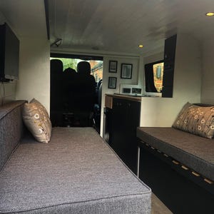May include: The interior of a van is converted into a living space with a grey couch bed, black cabinets, and a wood floor. The van has a white ceiling with wood paneling.