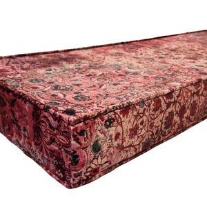 May include: A long, rectangular, red and pink patterned cushion with a floral design. The cushion is made of a soft, plush fabric and has a tufted top.