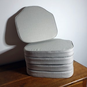 May include: A stack of four light grey chair cushions with rounded edges. The cushions are made of a soft, textured fabric.