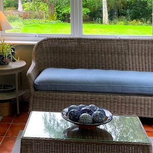 May include: A wicker sofa with a light blue cushion sits in front of a window with a view of a green lawn. A glass coffee table with a bowl of blue and white decorative balls sits in front of the sofa.