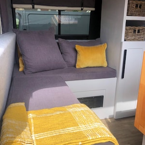 The interior of a renovated camper van with dark grey firm foam bench cushions inside. The converted campervan has comfortable thick upholstery foam cushions held in place with velcro strips on the underside of the cushions. The motor home bed cushions pull out to form a sleeping area with memory foam for comfort. The stain resistant bench cushion covers for the camper van conversion are made to measure for a plywood camper van build. Cut outs to fit around the van wall shaping for a perfect fit