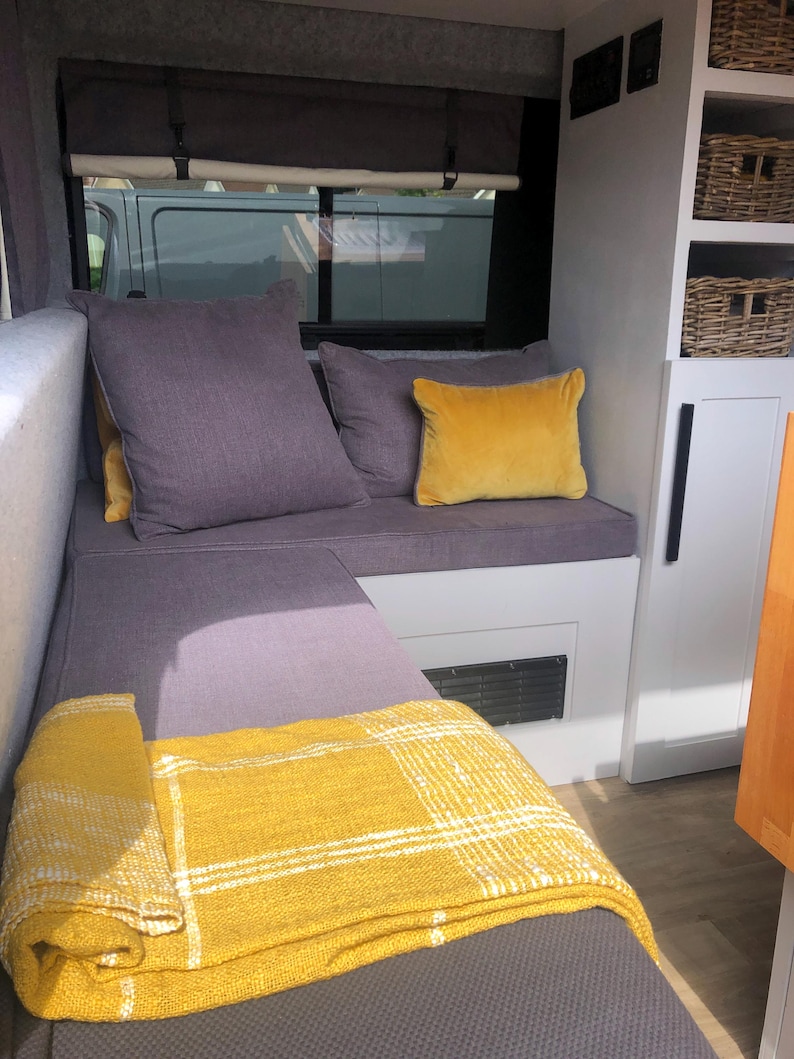The interior of a renovated camper van with dark grey firm foam bench cushions inside. The converted campervan has comfortable thick upholstery foam cushions held in place with velcro strips on the underside of the cushions. The motor home bed cushions pull out to form a sleeping area with memory foam for comfort. The stain resistant bench cushion covers for the camper van conversion are made to measure for a plywood camper van build. Cut outs to fit around the van wall shaping for a perfect fit