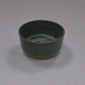 Peut inclure: Un petit bol rond en céramique présentant une glaçure vert forêt profonde sur l'intérieur et la partie supérieure extérieure, passant à une base en argile brune brute non émaillée. Le bol a une forme cylindrique simple et une finition mate.