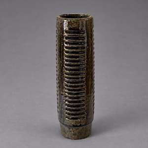 Pode incluir: Um vaso cilíndrico alto de cerâmica com um esmalte manchado de castanho escuro. O exterior apresenta um padrão vertical repetitivo de ranhuras horizontais rebaixadas que percorrem o comprimento do corpo, terminando numa base lisa.