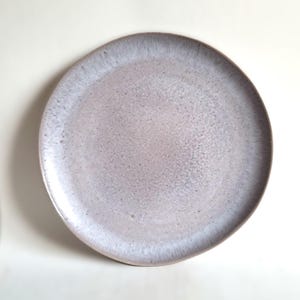 May include: A round, speckled ceramic plate with a light purple and white glaze. The plate has a slightly raised edge and a smooth, glossy finish. This plate is suitable for serving food.