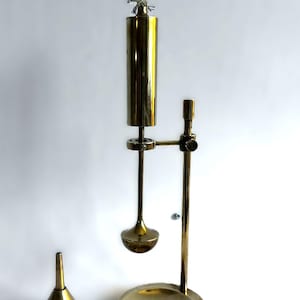 Puede incluir: Un aparato de laboratorio de latón antiguo con una parte superior cilíndrica, una base redonda y un embudo. El aparato tiene un soporte vertical con un brazo horizontal. El embudo es cónico.