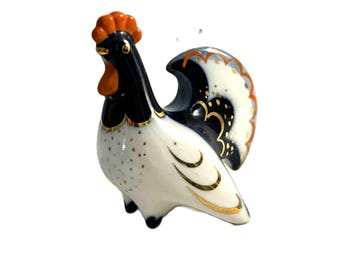 May include: A ceramic figurine of a rooster, featuring a white body with gold accents. The rooster has a black head and neck, an orange comb and wattle, and a detailed tail with black, gold, and white patterns. A decorative item.
