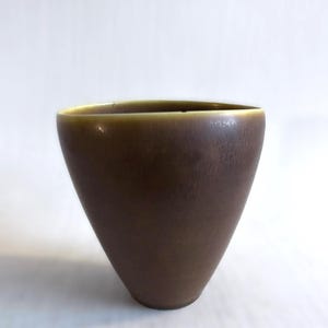 May include: A brown, cone-shaped ceramic vase with a slightly flared rim. The vase has a smooth, matte finish and a warm, earthy tone. The interior rim has a lighter, golden-brown color. The vase is empty and sits on a white surface.