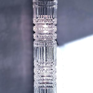 Puede incluir: Una lámpara de vidrio transparente y vertical con base y parte superior negras. La lámpara tiene un diseño geométrico texturizado con múltiples fuentes de luz visibles dentro del vidrio. La lámpara mide aproximadamente 30 cm de alto.